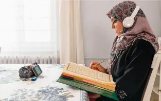 Older Muslim woman sitting on chair taking online Islamic courses for ladies on mobile while writing notes in Quran book in USA and UK
